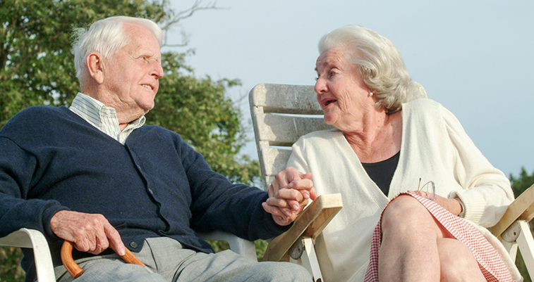 Äldre man och kvinna sitter utomhus i varsin stol. Båda är glada och tittar på varandra.