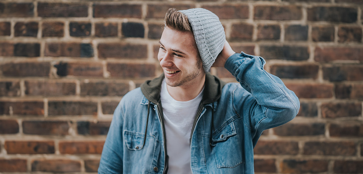 glad kille med mössa och jeansjacka mot tegelvägg