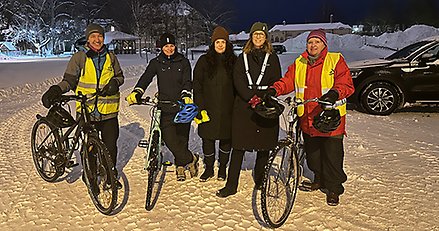 fem personer står med reflexvästar och cyklar på en snöig parkering
