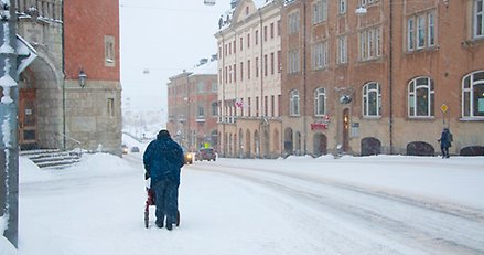 En person går med rollator på en trottoar i en stadsmiljö och det snöar ymnigt.